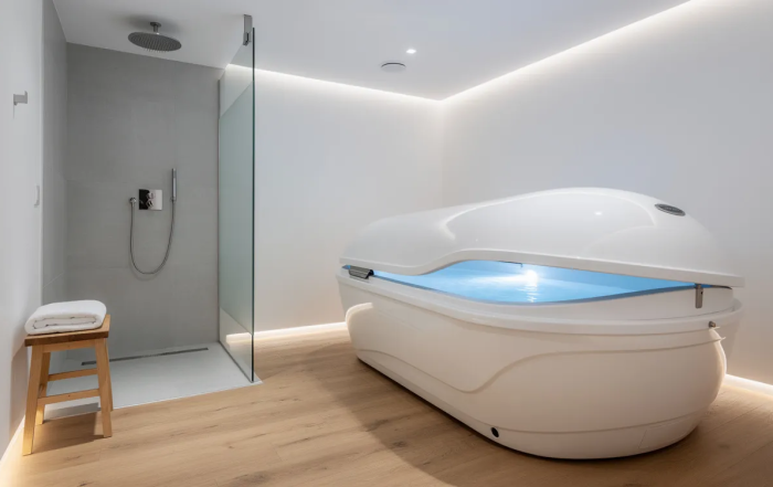 The image depicts a modern float pod situated in a clean, minimalist private room, illuminated by soft ambient lighting, with a small shower area nearby. This serene environment is designed for float therapy, promoting deep relaxation and sensory deprivation, allowing individuals to experience a unique zero gravity environment that aids in stress relief and overall well-being.